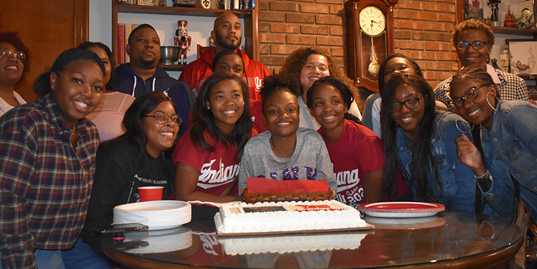 A big group of students smiling behind a cake. 