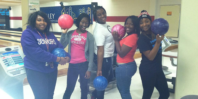 Five students in a bowling alley posing with their bowling balls. 