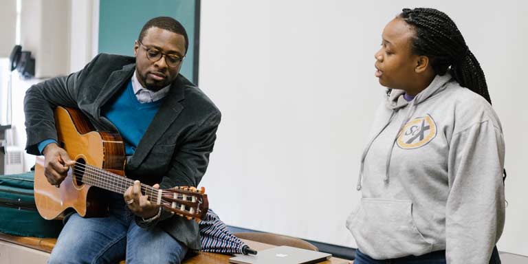 A professor playing the guitar while a student sings. 