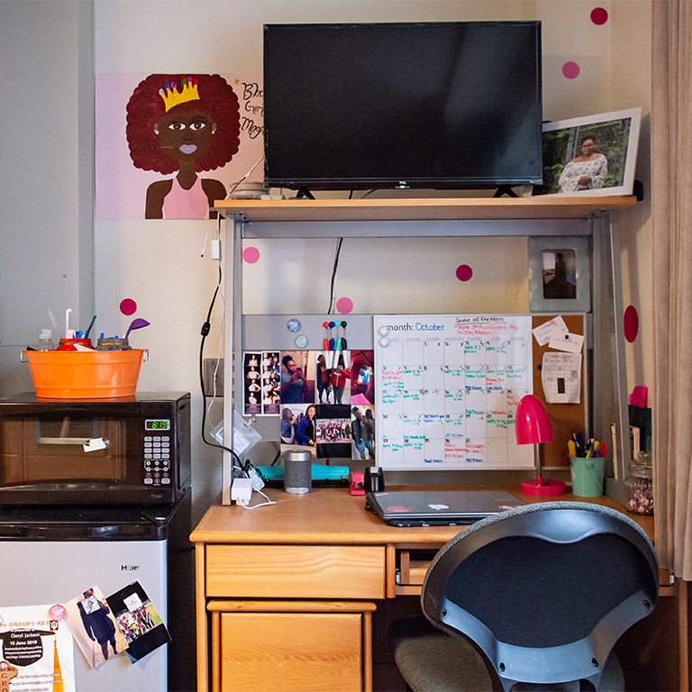 A student's desk with a television above it and a mini fridge and microwave beside it. 
